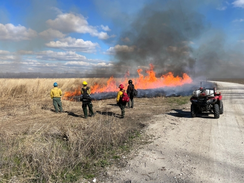 A large fire burns through a grassland as four people in safety gear stand watching.