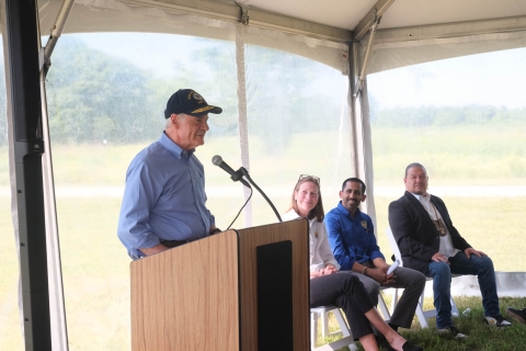 a man wearing a ballcap stands speaking into a podium microphone while three people sit and listen