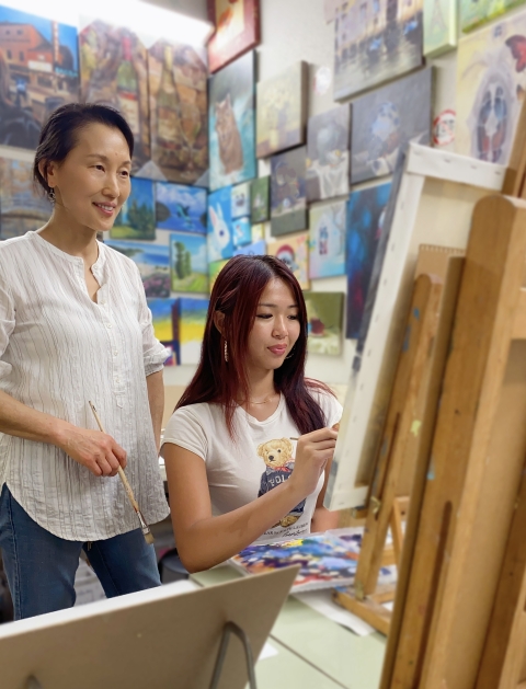 Young artist sits at easel in studio with teacher standing behind.