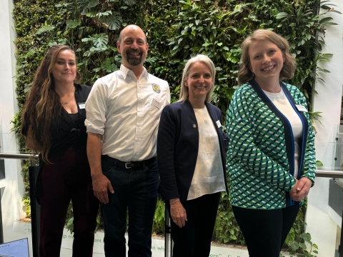 Four people stand in a line smiling for the camera. From left to right: a woman wearing a black pantsuit with long brown hair, a man wearing a white FWS-branded button up, a woman with blonde hair in a blue blazer and white shirt, and a woman with short blonde hair and a green and blue patterned cardigan over a white shirt. 