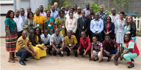 A group of approximately 30 people line up for a group photograph, many are wearing colorful clothes with traditional African prints. 