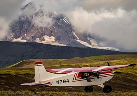 A fixed wing wheeled aircraft with red wings and tail and white body on tundra with rising hills and mountains in the distance..