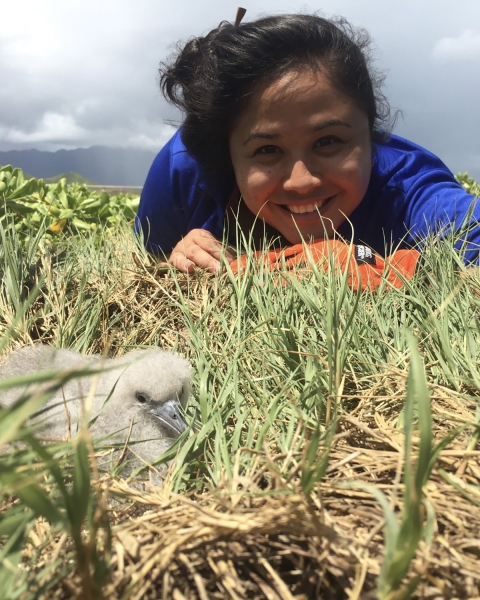 Smiling person in blue shirt lays on ground next to seabird chick in grass. 