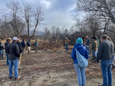 A group of people stand in a circle and talk. Trees and mountains are in the distance.