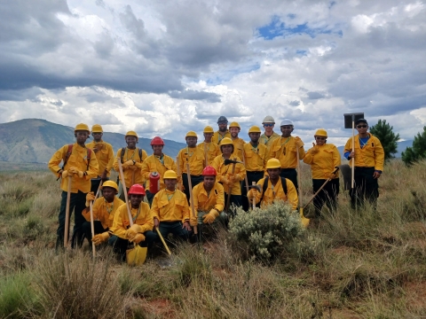 A group of firefighters stand together in their Nomex and post for a photo as a group. The landscape is dry vegetation and there are clouds in blue sky.