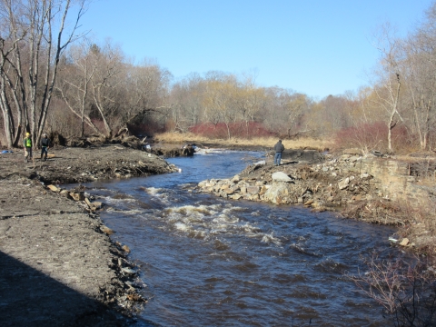 a river flows, with gray rock and soil on the left and debris from a former dam on the right. bare trees and blue sky are in the background.
