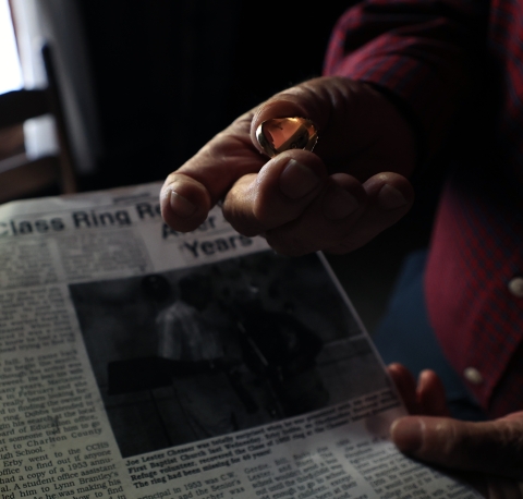 Close up of Joe's hand holding the class ring he lost while bringing in wood for the fire and found by a refuge visitor. A newspaper article about finding the ring is in the background.