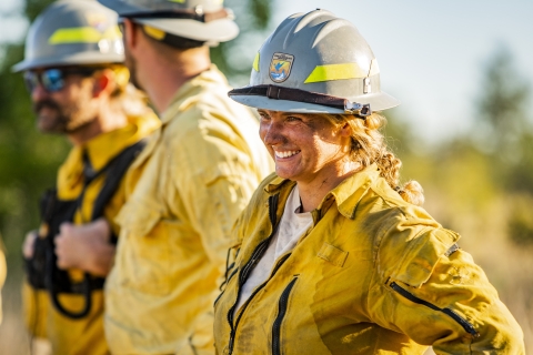 A firefighter smiles as a photo is taken of her. There are two other firefighters in the background that are blurry.