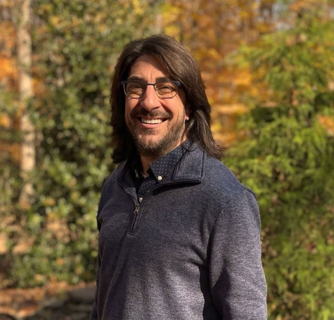 A person standing against a background of trees wearing glasses, and smiling. 