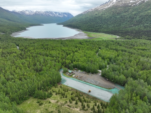 Aerial drone image of Eklutna Lake ATV trailhead parking lot with the lake in the background. Forest surrounds the area and snow topped mountains on the horizon. 