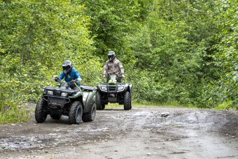 Two all terrain vehicles with riders wearing helmets on a muddy off highway vehicle trail in a wooded area. 