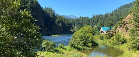 A landscape view of a river surrounded by trees and mountains. A building with a light blue roof sits nestled within the landscape. 