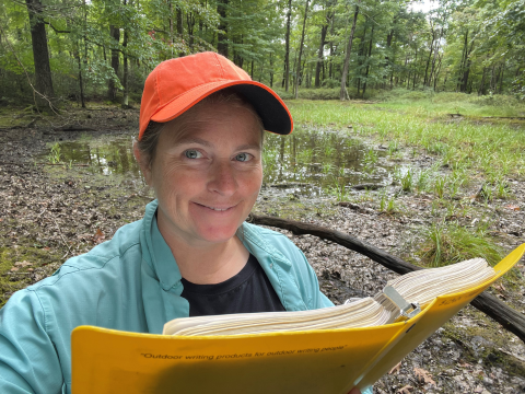 A selfie of a woman holding a notebook standing near a wetland in a forest