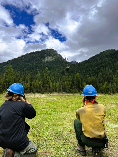 In a mountainous environment two firefighters kneel and observe their coworkers drop from a line suspended below a helicopter. 