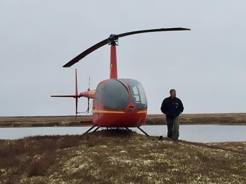 A person stands next to a parked helicopter on a tundra knoll with a river and shore behind them.