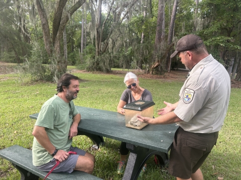 A male Fish and Wildlife biologist talking to two people sitting at a green picnic table while they look at a terrarium 