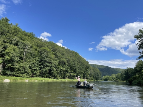 2 people in a boat on a waterbody surrounded by trees with a mountain in the background