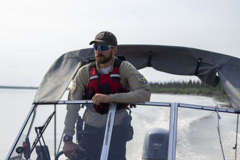 A view from the bow of a boat towards the stern of a Federal Wildlife Officer at the helm with the Yukon River in the background.