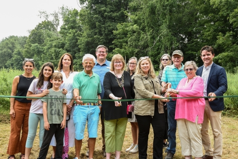 a large group of people stands behind a green rope, with green grass and trees in the background