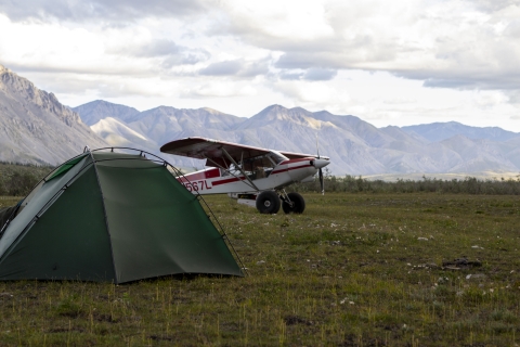 A small tent with a Top Cub bush plane parked behind it on a grass runway area with mountains in the background.