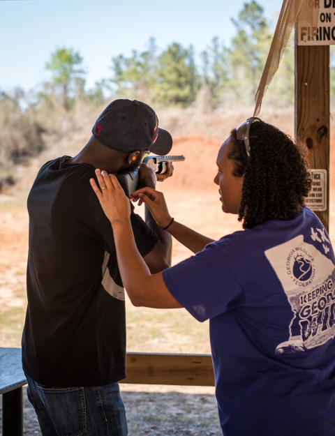 Mentor shows student correct sporting arms stance and stock placement while student aims at target.