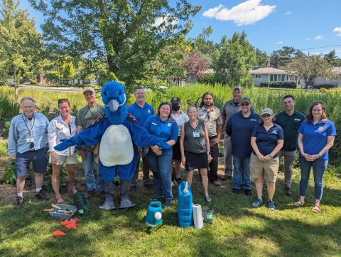 group of people standing with a person in a blue and white goose mascot costume in a shaded garden patch