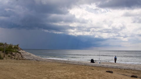 A person in waders stands on the beach fishing.