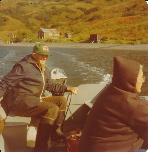 Kristjan Helgason operates the motor on the back of a skiff boat on water and Clara Helgason wears a hooded sweatshirt sitting facing the side of the boat. Their camp of buildings sit at the foot of a hill.