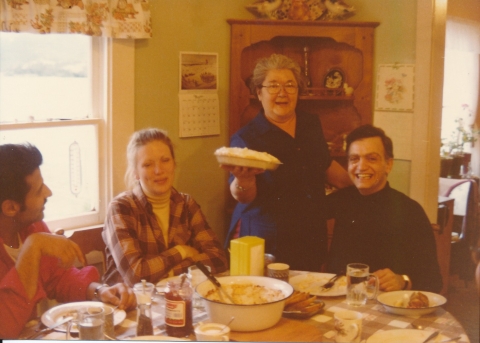 Three people sit at a dining room table with plates and dishes on it. Clara Helgason holds a pie and smiles.