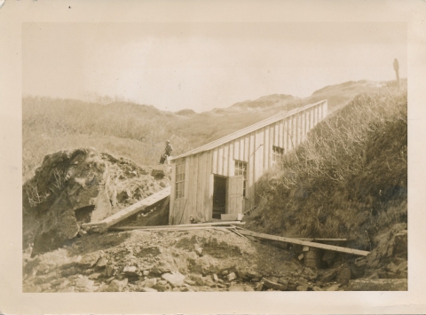 A wood building with a slanted roof with planks stretching across rugged dirt. sloped hills rise beside the building and a person walks on the hill behind the building. A mountain rises behind them.