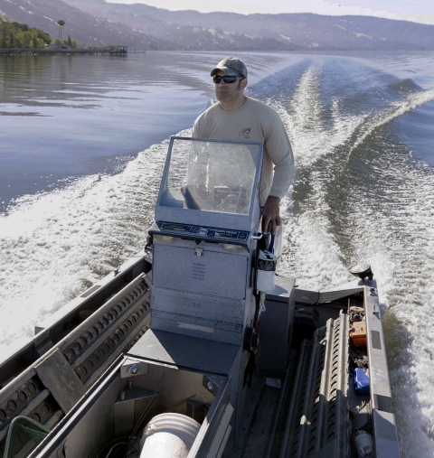 Man wearing sunglasses and hat in boat speeding along a lake.