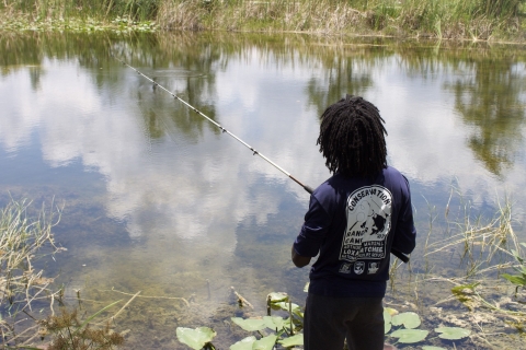 student holds a fishing pole facing a lily-filled pond; back of T-shirt says “Conservation Ranger Camp [year obscured] Arthur R. Marshall Loxahatchee National Wildlife Refuge”