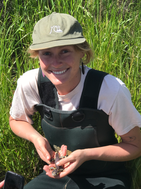 A woman wearing a hat and overalls, smiling as she holds a small salamander in her hands