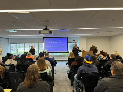 Uniformed federal wildlife inspector stands in front of seated audience with presentation on screen behind them and two other uniformed staff assist