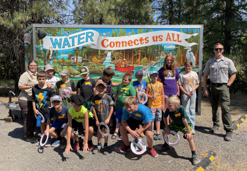 Several kids pose in front of a colorful mural with a banner that says, "Water Connects us ALL"