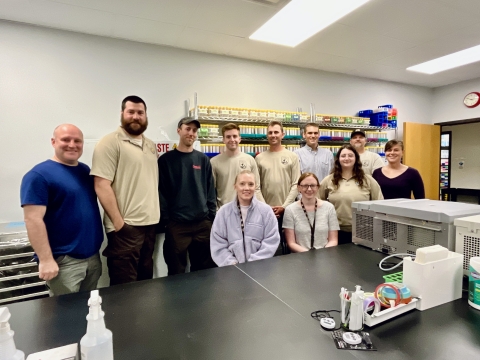 11 people stand behind lab counter with a shelf of sample tubes in the background