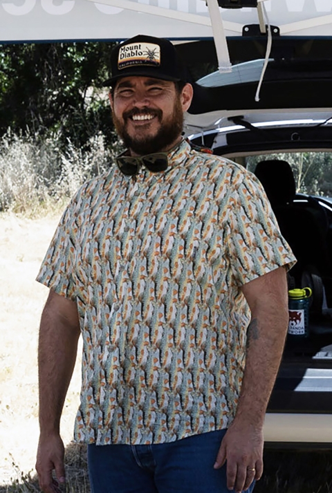 Man with black hat "Mount Diablo" with sunglasses around next stand under a tent with bushes and van in background