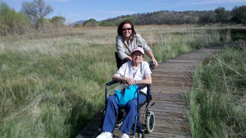 2 people pose for photo, one older, in wheelchair on boardwalk through grass