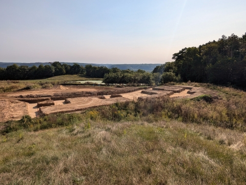 exposed dirt at building site that is surrounded by green trees in the distance and dry grasses in foreground