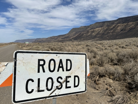 On road barrier, the image is up close to a sign stating "Road Closed". In the background is a sage step with portions burned from wildfire.