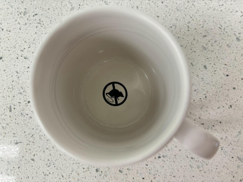 An empty mug with a printed ink silhouette of a rat inside a circle with a line through it at the bottom of a mug.