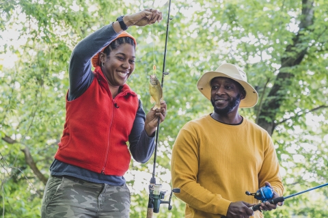 woman reels in small fish while man watches intently 