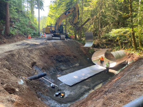 A construction site in the forest with a deep dirt pit with corrugated sheeting and workers wearing safety vests and hard hats. Heavy equipment and trees in the background.