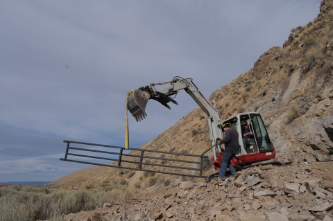 A mini track ho holds a metal grate in the air. A person is driving the track ho, and a man is standing next to it. 