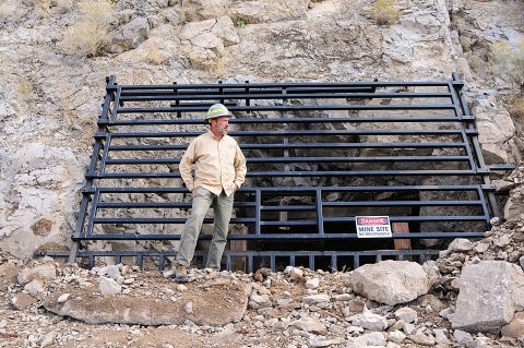 A man in a yellow shirt with a hard hat on stands by a grate covering an opening in the rocks. He is looking over his left shoulder.