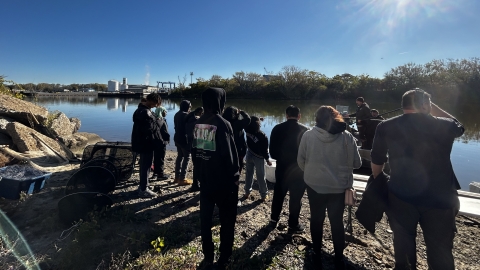 Image of students with teacher gathered along the shore with boat.