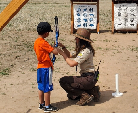 A person in USFWS uniform showing a child how to do archery. In the background, there are targets.