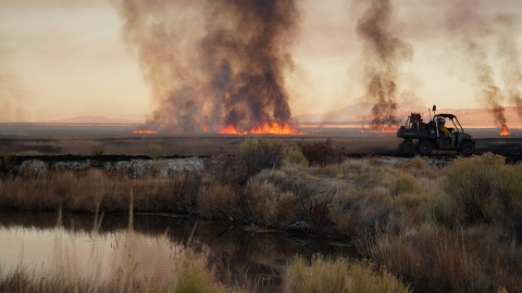 Wildland firefighters patrol a prescribed fire by UTV. Islands of smoke and flames dot the background of the Great Basin high desert landscape. 