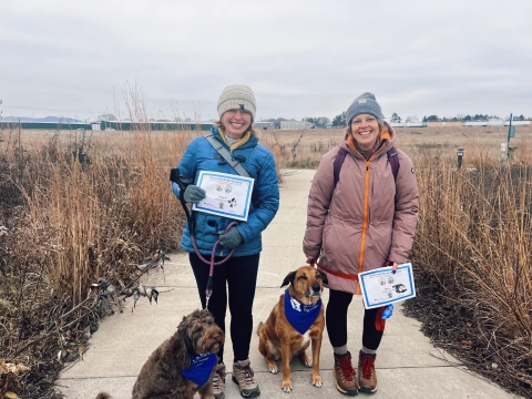 Two smiling visitors pose holding their BARK Ranger certificates next to their leashed dogs wearing BARK Ranger bandanas