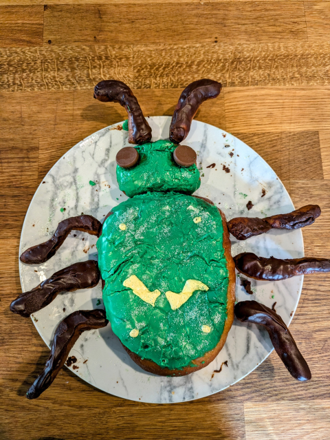 A large donut sitting on a plate. It is shaped like a beetle and has a green sparkly head and torso, and brown antenna, legs, and eyes